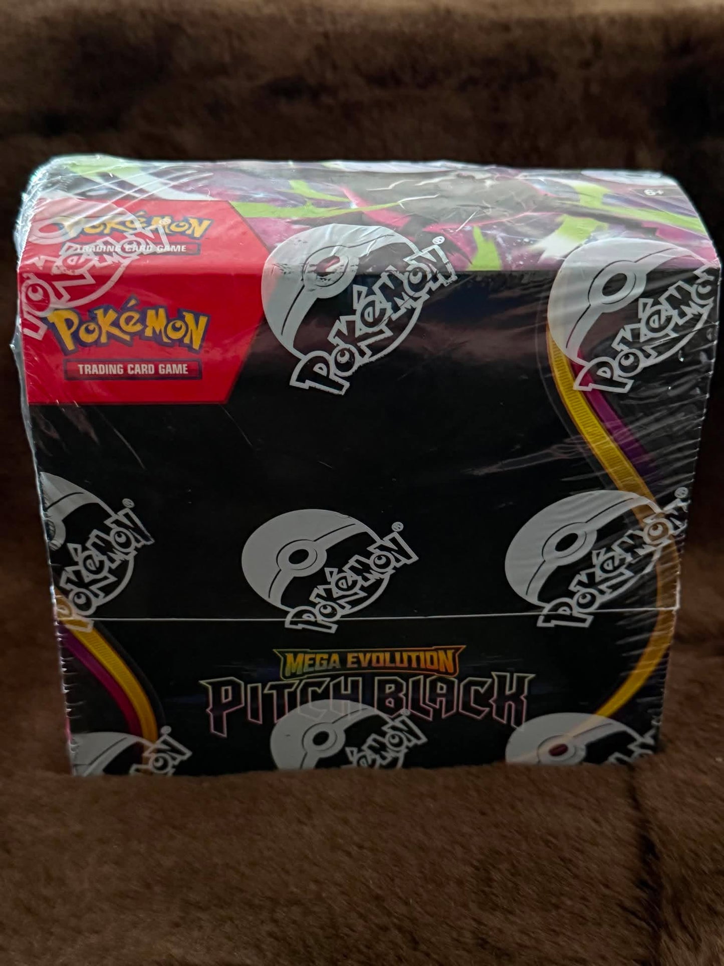 A sealed Pokémon Mega Evolution Pitchblack trading card game booster box wrapped in clear plastic, photographed on a brown textured surface.