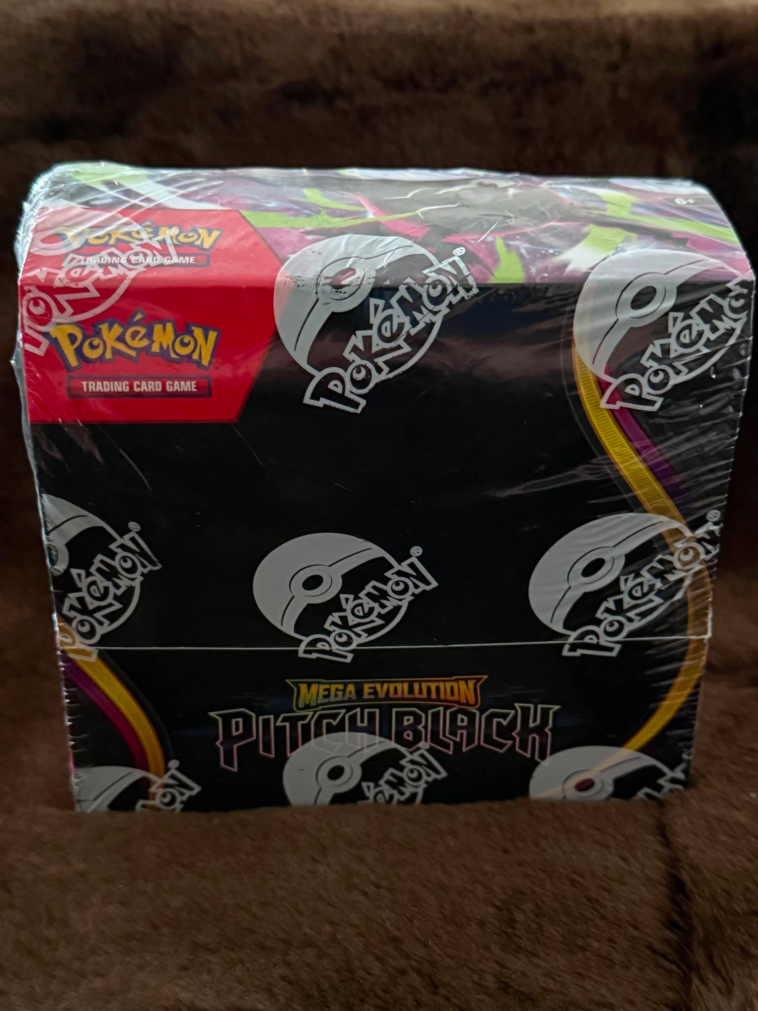 A sealed Pokémon Mega Evolution Pitchblack trading card game booster box wrapped in clear plastic, photographed on a brown textured surface.